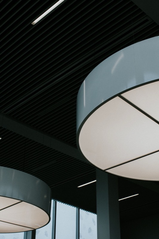 Modern drum-style commercial ceiling light fixtures against a dark slatted office ceiling